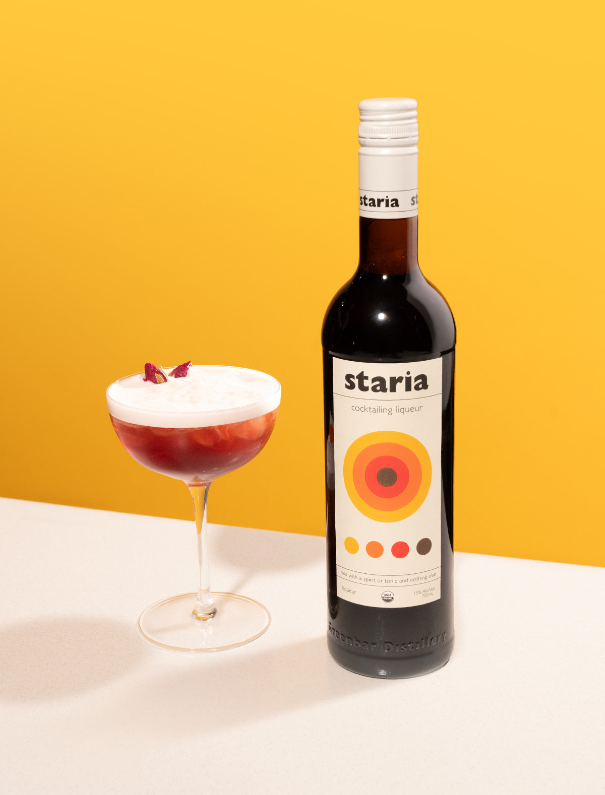 A bottle of Staria cocktail liqueur beside a red cocktail with a foamy top and floral garnish in a coupe glass, on a white table with a yellow background.