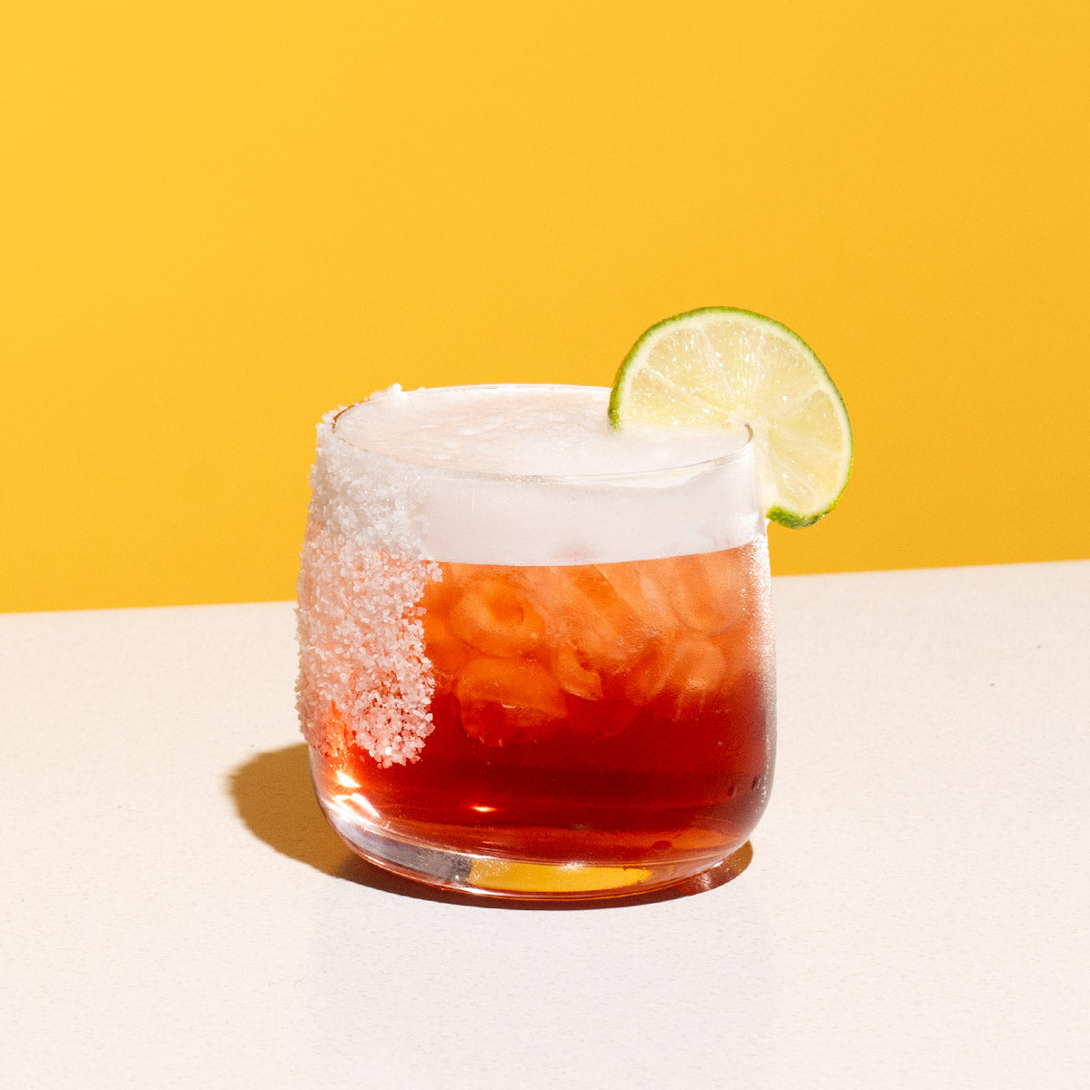 A red cocktail with a foamy top served in a rocks glass, garnished with a lime wheel and a salt rim, on a white table with a yellow background.