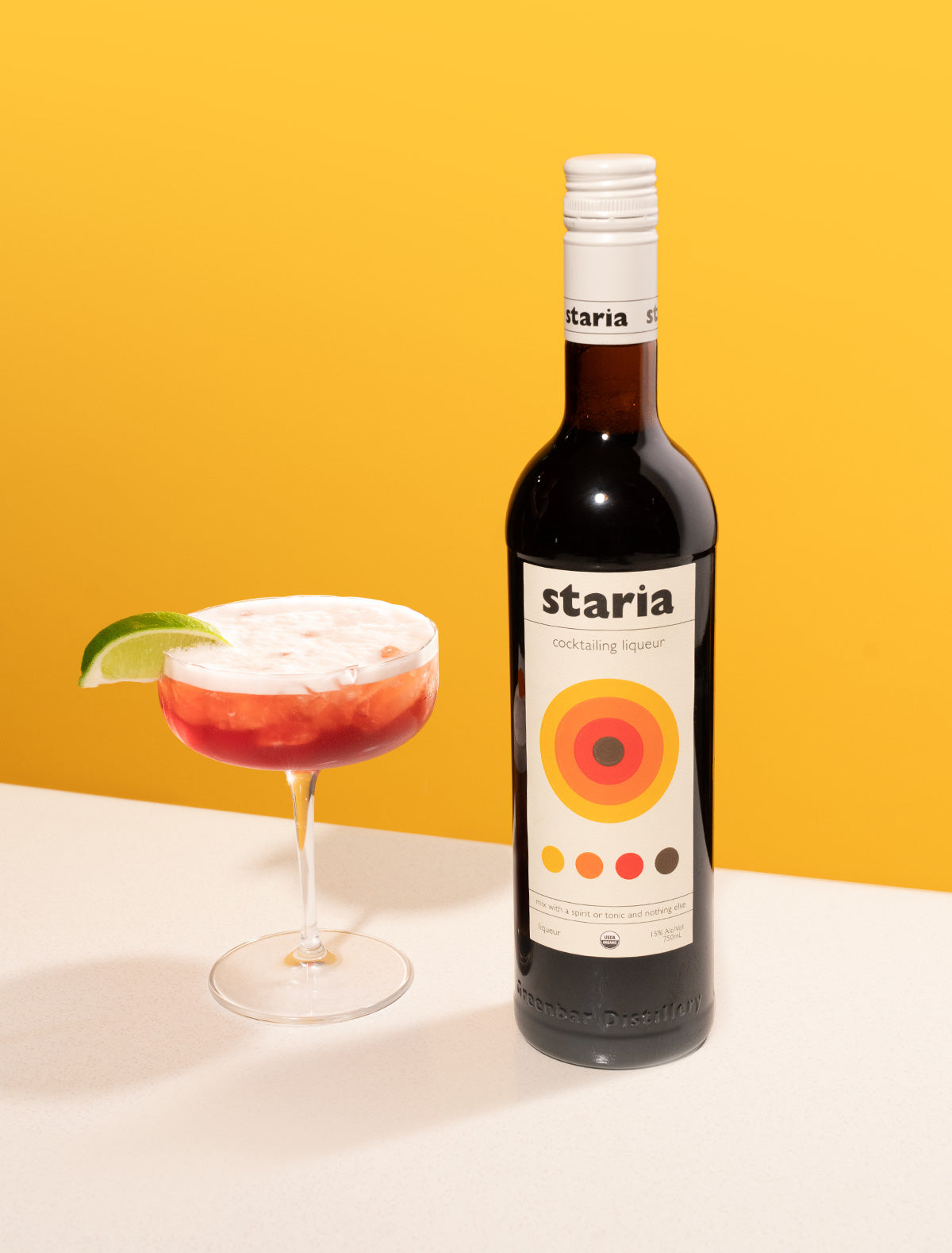 
A bottle of Staria cocktail liqueur next to a red cocktail with a foamy top and lime garnish in a coupe glass, set on a white table with a vibrant yellow background.