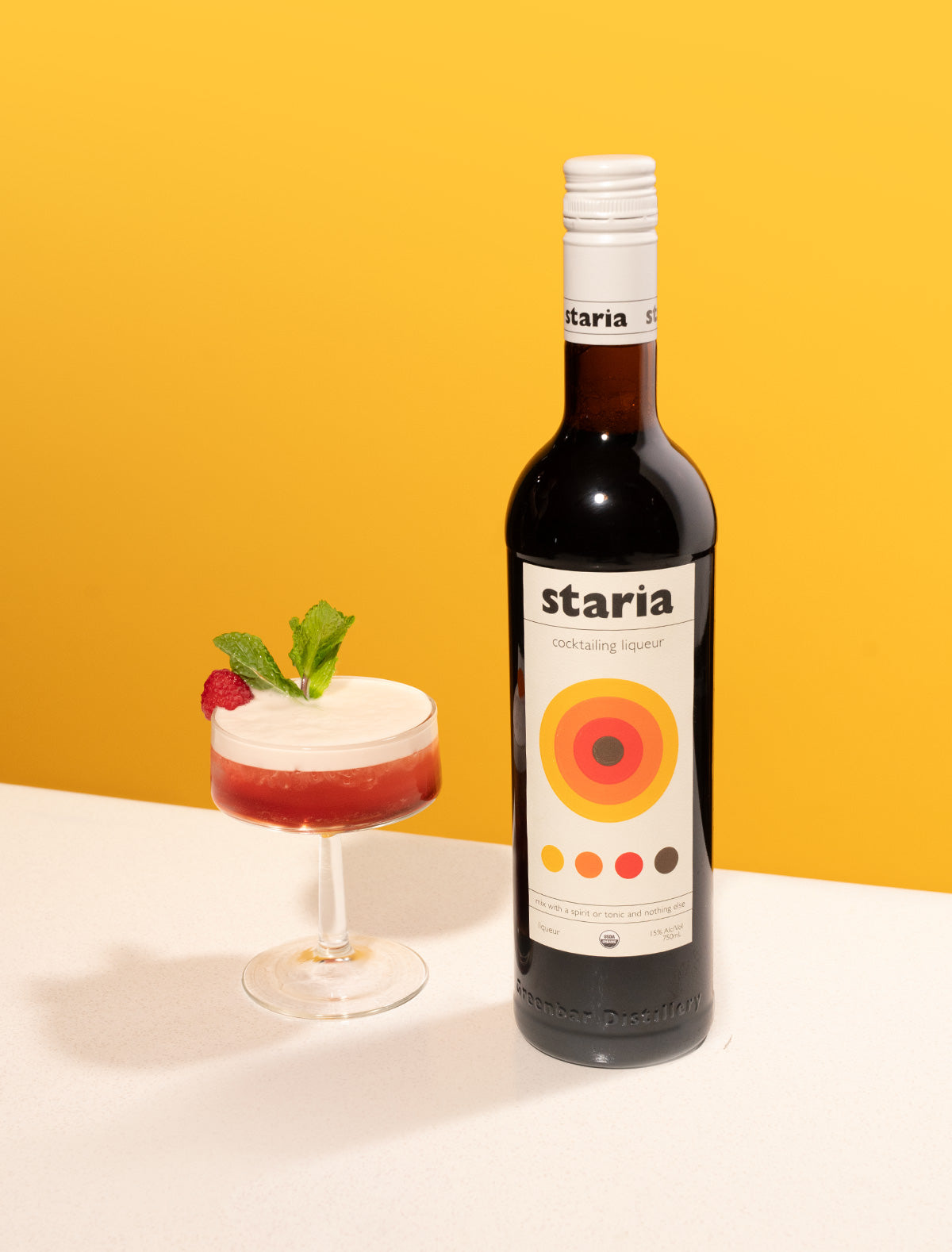 A bottle of Staria cocktail liqueur next to a red cocktail with a foamy top, garnished with a raspberry and mint sprig, on a white table with a yellow background.