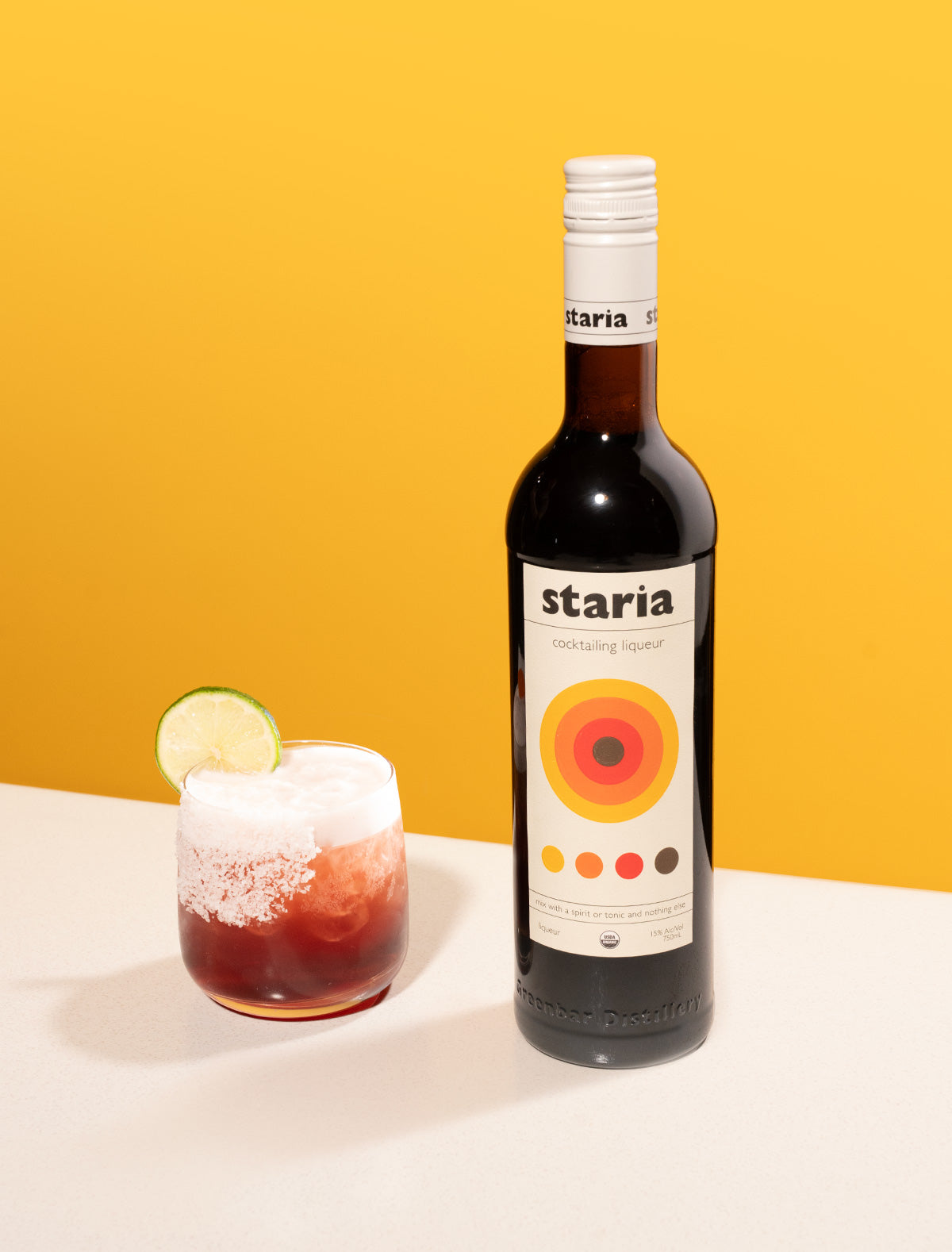 A bottle of Staria cocktail liqueur next to a red cocktail with a salt rim, lime garnish, and foamy top, on a white table with a bold yellow background.