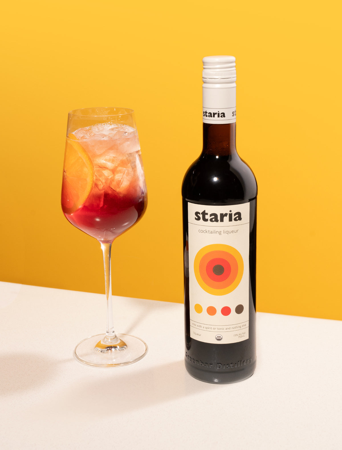 A bottle of Staria cocktail liqueur beside a red spritz cocktail with ice and an orange slice in a stemmed glass, set on a white table with a vibrant yellow background.