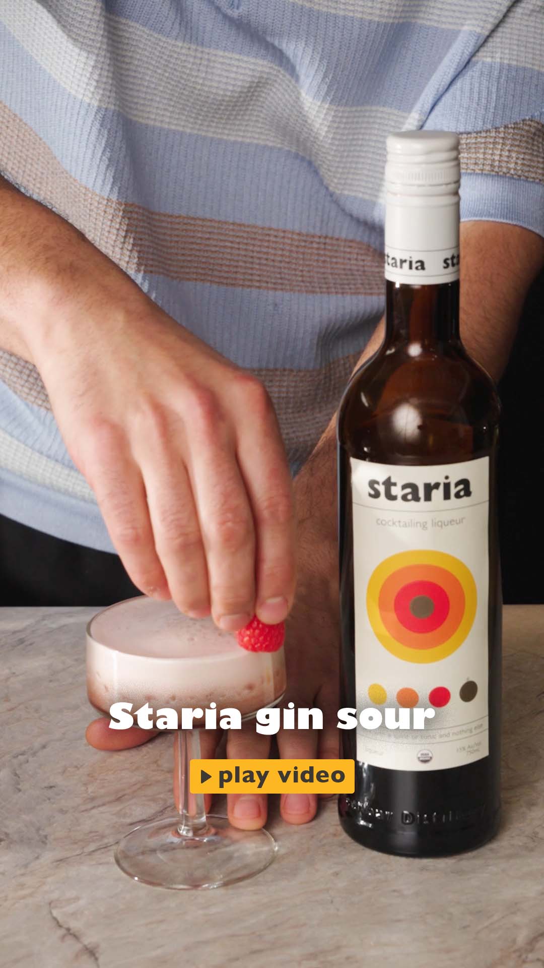 A man in a kitchen has a bottle of Staria in the right of the table and a bottle of Gin on the left, makes a gin sour by mixing Staria and gin in a shaker, straining it into a coupe glass, garnishing with mint and a raspberry, and smiling at the camera with the finished drink.