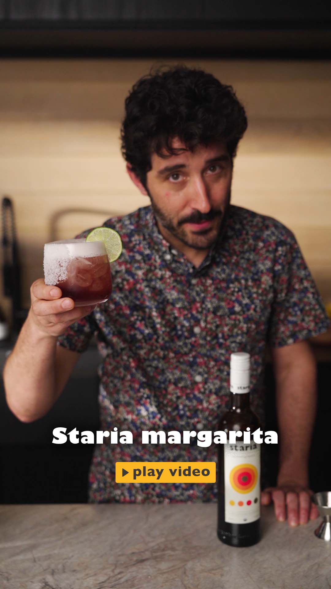 A man in a bright kitchen pours Staria and tequila into a shaker, mixes and strains a red margarita into a salt-rimmed glass, then garnishes it with a lime wheel and lifts it to the camera with a smile.