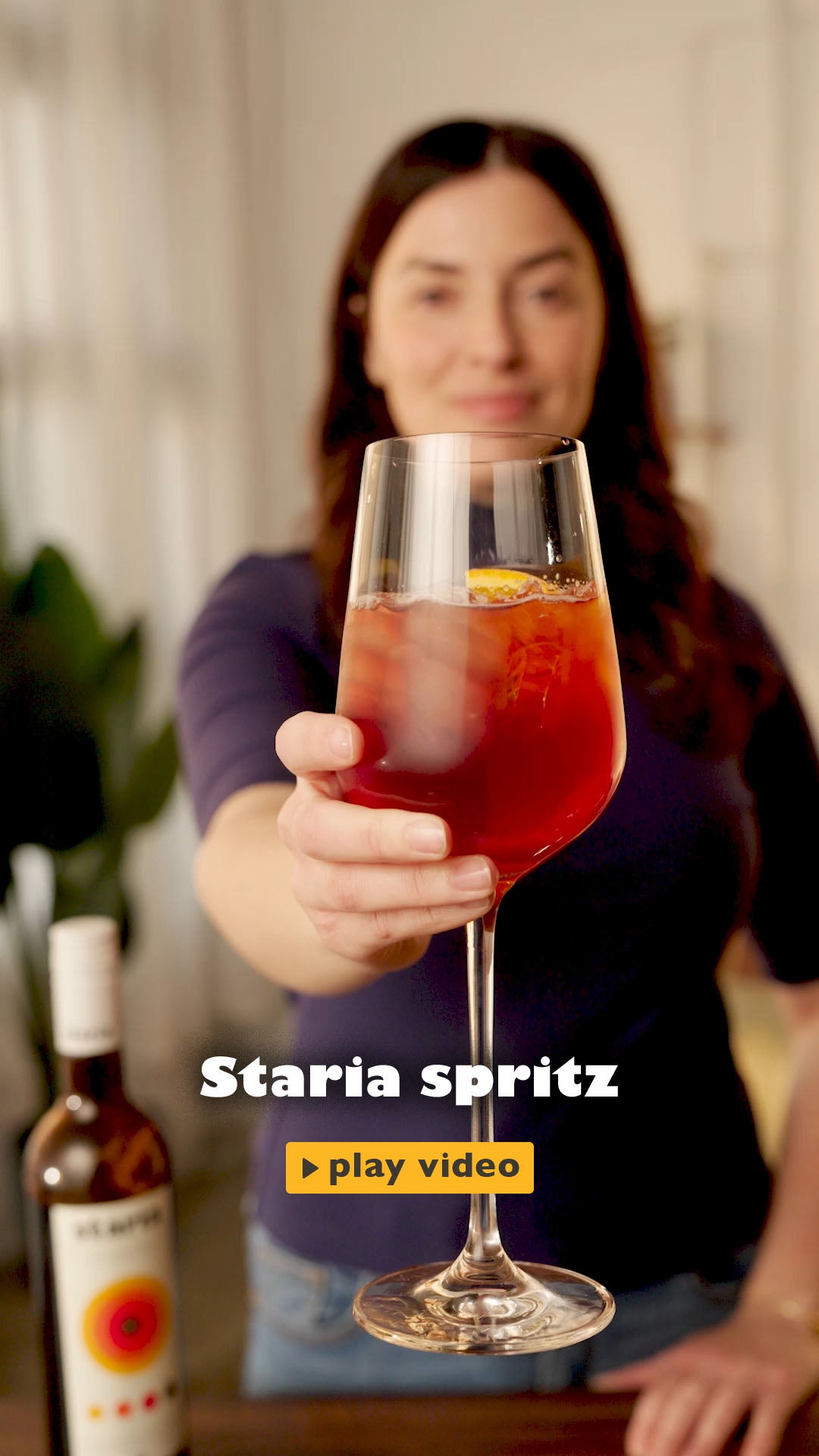 A smiling woman holds up a bottle of Staria cocktail liqueur, starts by picking up a stemmed wine glass filled with ice. Using a jigger, she pours in a measure of Staria, followed by a splash of sparkling water, then tops it with Prosecc,o then adds a slice of orange into the drink