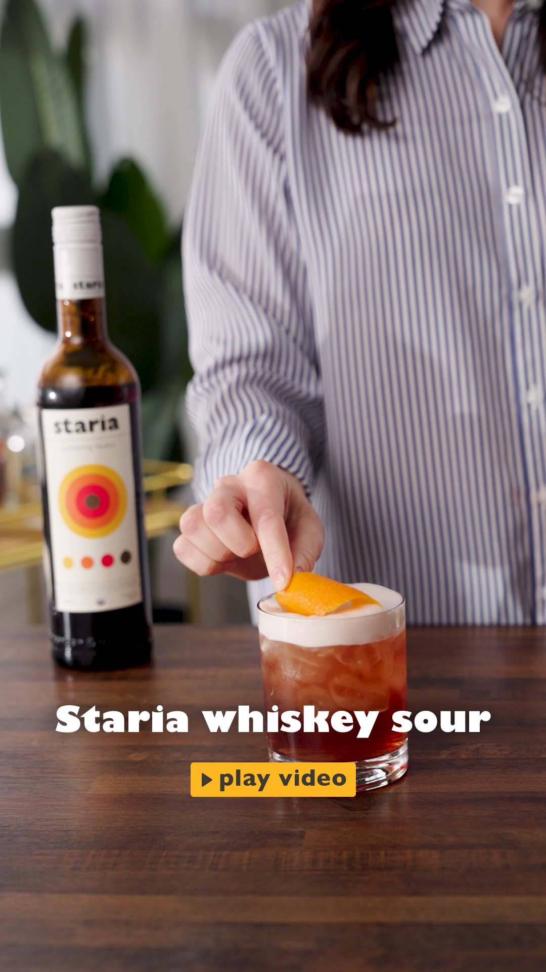 In a warmly lit home kitchen, a cheerful influencer walks up to the counter holding a bottle of Staria cocktail liqueur. They carefully measure out a shot using a jigger, pouring it into a cocktail shaker. A bottle of bourbon follows, adding depth to the drink. After sealing the shaker, they give it a firm, rhythmic shake. The mixture is then strained into a rocks glass filled with ice, revealing a rich, reddish drink with a frothy top.  Final touch, she twist an orange peel and place it neatly on the rim