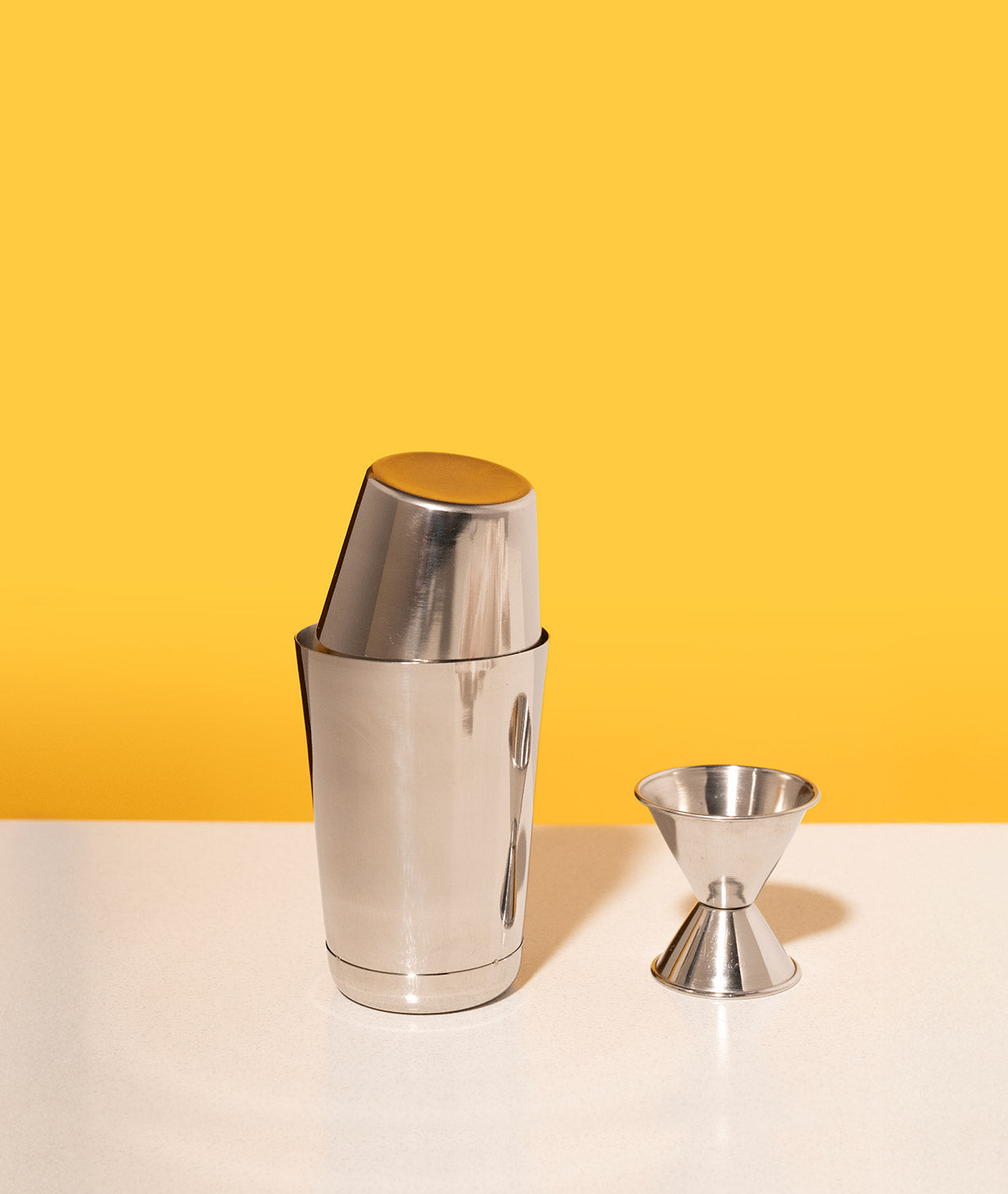 A stainless steel cocktail shaker and jigger on a white table with a vibrant yellow background.