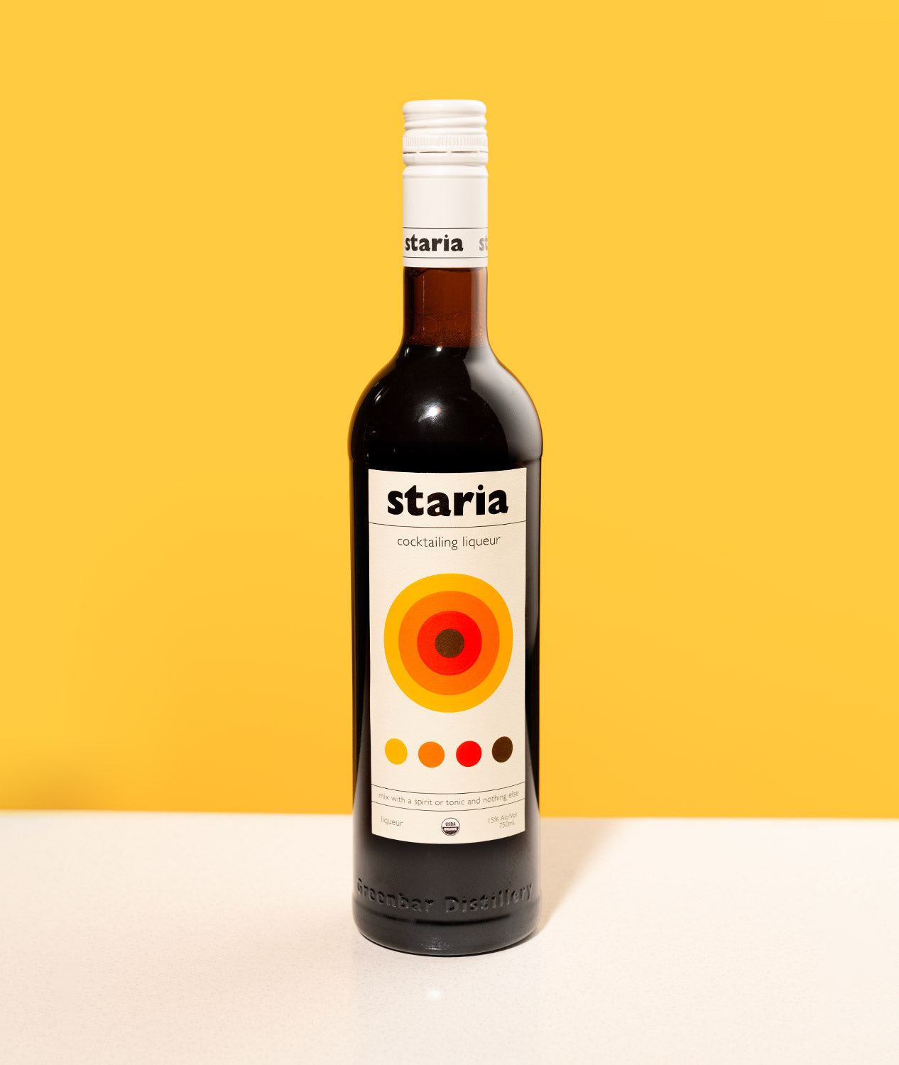 A bottle of Staria cocktail liqueur standing alone on a white table with a bold yellow background.