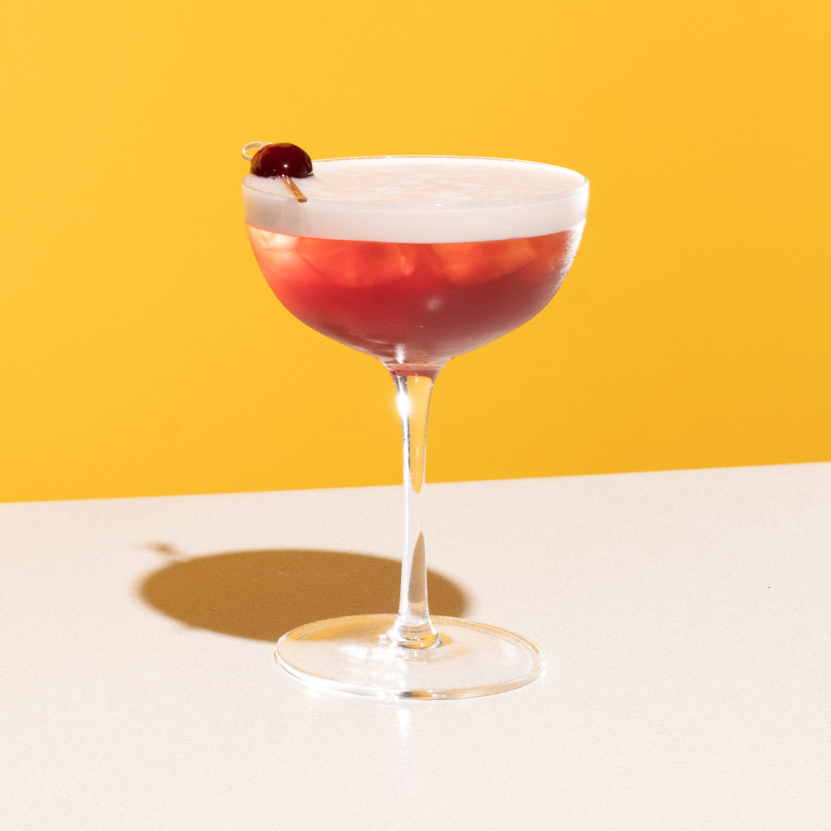 A red cocktail with a foamy top served in a coupe glass, garnished with a cherry on a cocktail pick, on a white table with a yellow background.