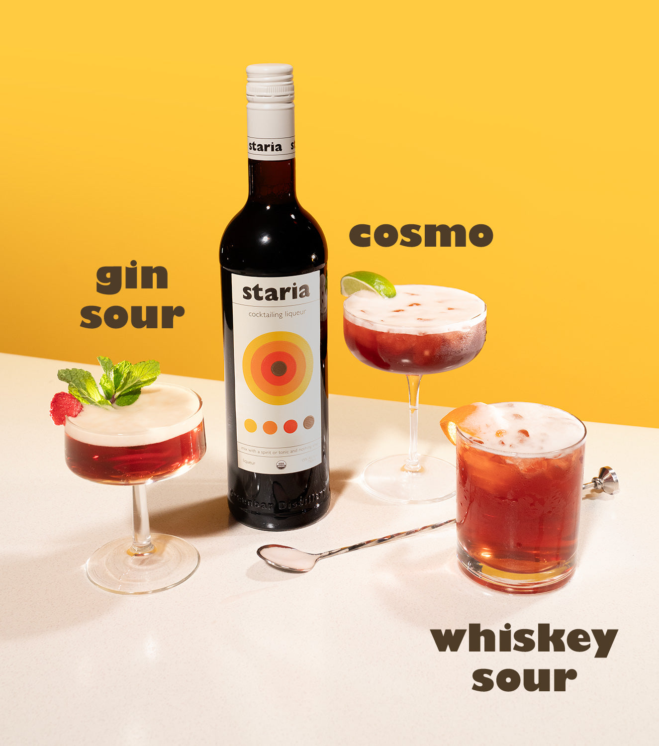 Three colorful cocktails—gin sour, cosmo, and whiskey sour—surround a bottle of Staria cocktail liqueur on a white table with a bold yellow background. Each drink is garnished and labeled with playful typography.